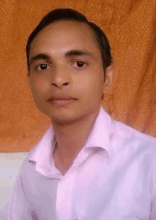 a young man in a pink shirt is sitting in front of an orange curtain and looking at the camera .