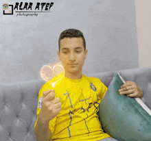 a young man in a yellow shirt is sitting on a couch and holding a heart in his hand