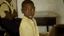 a young boy in a yellow shirt is sitting at a desk in a classroom making a funny face .