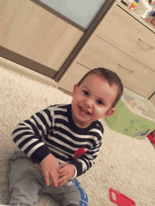 a little boy wearing a striped shirt is sitting on the floor and smiling