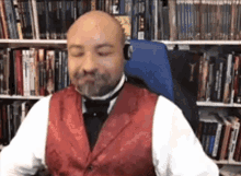 a bald man with a beard wearing a red vest and bow tie is sitting in a chair in front of a bookshelf .