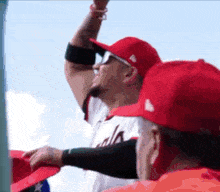 a baseball player wearing a red hat and sunglasses stands in the stands with his arm in the air .