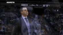 a man in a suit and tie stands in front of a crowd at a basketball game with #shaotin written on the bottom