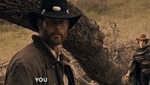 a man in a cowboy hat is standing next to a tree with the words " you " written on his jacket