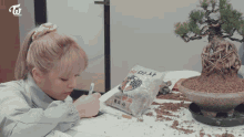 a woman sits at a table with a bonsai tree and a bag of dirt with the letters twice on it