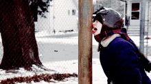 a person wearing a helmet and scarf is sticking their head through a chain link fence in the snow .