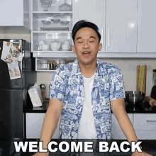 a man in a blue and white shirt is standing in a kitchen with the words welcome back below him