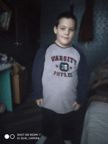 a boy wearing a varsity physics shirt stands in a dark room