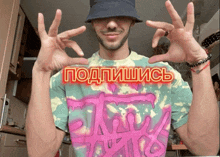 a man wearing a tie dye shirt and a bucket hat holds up his hands in front of a sign that says подписаться