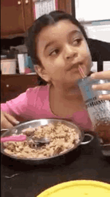 a little girl is drinking milk through a straw while sitting at a table with a bowl of cereal .