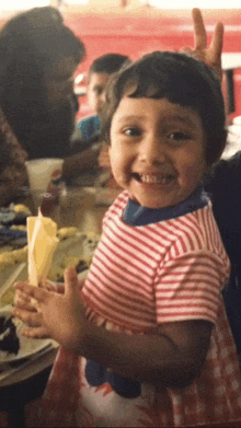 a little girl in a red and white striped shirt is smiling and giving a peace sign