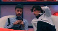 two men are sitting next to each other on a couch and one of them is looking at his phone .