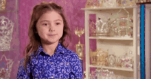 a little girl in a blue shirt is standing in front of a shelf filled with trophies and tiaras .