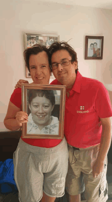 a man in a red england shirt holds a framed photo of a woman