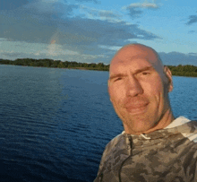 a bald man stands in front of a lake with a rainbow in the background