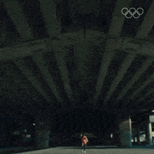 a man in an orange tank top and black shorts is running under a bridge with the olympic rings in the background