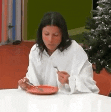 a woman in a white robe is sitting at a table eating a plate of food with a fork and knife .