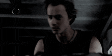 a man in a black tank top is sitting in a dark room and looking at something .