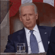 a man in a suit and tie is sitting in front of an american flag with a glass of water .