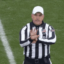 a referee wearing a black and white striped shirt and a white hat is standing on a field with his hands crossed .