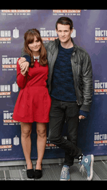 a woman in a red dress and a man in a black jacket pose in front of a doctor sign