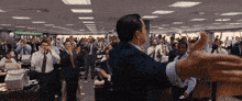 a man in a suit stands in front of a crowd of people in an office with a sign on the wall that says " stocks "