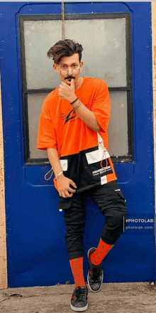 a man wearing an orange t-shirt and black pants stands in front of a blue door