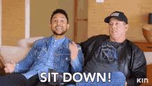 two men are sitting on a couch and one of them says " sit down "