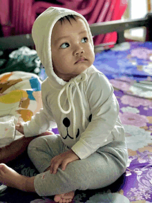 a baby wearing a white hoodie and gray pants sits on a purple bed