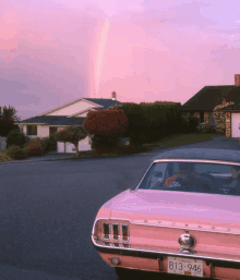 a pink mustang is parked in front of a house with a rainbow in the background