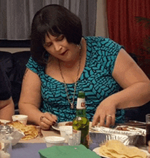 a woman in a blue shirt is sitting at a table with food and a bottle of beer