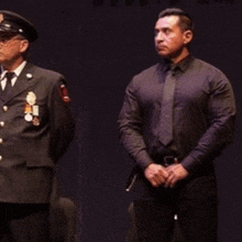 a man in a black shirt and tie stands next to another man in a uniform