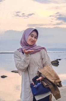 a woman in a hijab is standing on a beach holding a cell phone and a jacket .