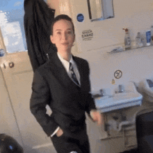 a woman in a suit stands in front of a bathroom sink with a no smoking sign on the wall