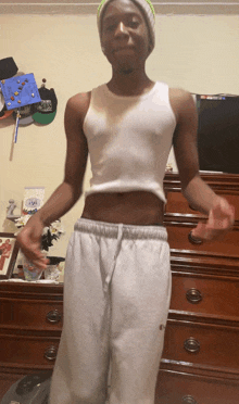 a young man wearing a white tank top and grey sweatpants is standing in front of a dresser