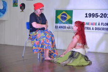 a woman sits on a chair next to a woman kneeling down in front of a sign that says 1995-20