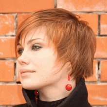 a woman wearing short red hair and red earrings stands in front of a brick wall