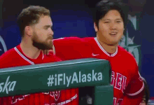 two baseball players are talking to each other in a dugout with a sign that says ifly alaska on it .