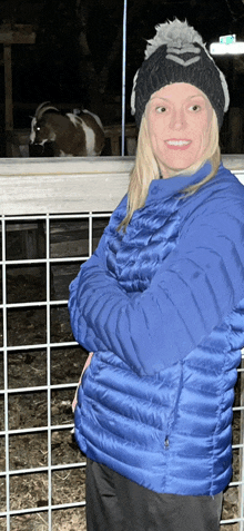 a woman wearing a blue jacket and a black hat stands in front of a goat