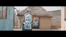 a man wearing a blue and white tank top with a snake on it stands in front of a thatched hut