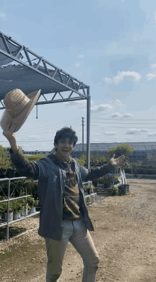 a man holding a straw hat in the air in front of a greenhouse