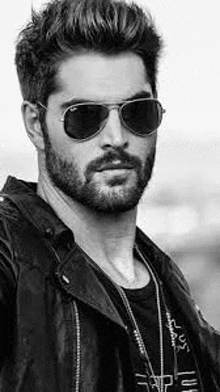 a man with a beard and sunglasses is wearing a leather jacket and a necklace .