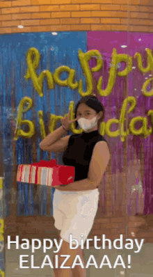 a woman wearing a mask is holding a gift box in front of a wall with balloons that say happy birthday .