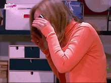 a woman in an orange sweater covering her face with her hands in front of a shelf with books on it