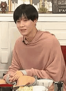 a young man in a pink sweater is sitting at a table with food .