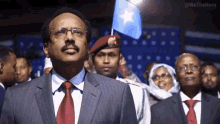 a man in a suit and tie stands in front of a crowd holding a blue flag with a white star on it