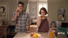 a man and a woman drinking orange juice in a kitchen with a bottle of orange juice in the foreground