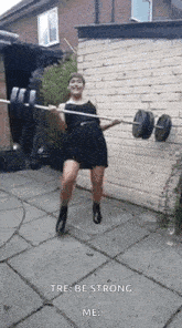 a woman in a black dress is holding a barbell in her hands in front of a brick wall .