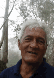 a man with gray hair and a blue shirt is smiling in front of trees