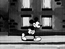 a black and white cartoon of mickey mouse walking down a sidewalk in front of a building .